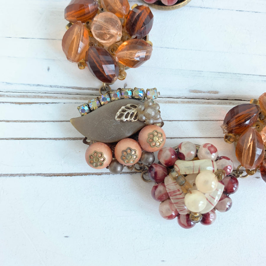 A close-up of a brown, gray, and white beaded necklace. The necklace is made of various shapes and sizes of beads, including round, oval, and rectangular beads. The beads are strung together on a metal chain. The necklace is lying on a white wooden surface.