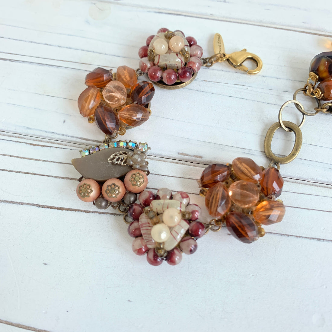 A brown and gold beaded bracelet with a variety of beads including clear, brown, and white beads. The bracelet has a metal clasp and a small metal charm with a leaf design.