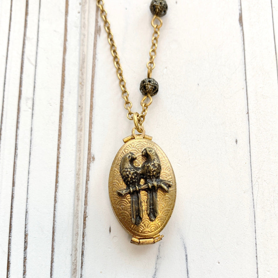 Gold oval locket necklace on a gold chain. The opening locket is embellished with a bronze stamping of two birds on a branch.