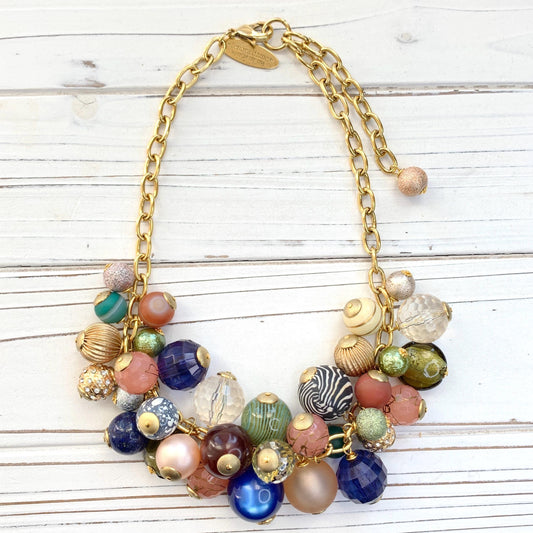 May include: Gold chain necklace featuring a cluster of colorful beads. Beads include various textures and patterns:  clear, frosted, glittery, striped, and solid colors such as peach, teal, green, blue, brown, and cream.  The necklace has a gold clasp. Lenora Dame necklace.