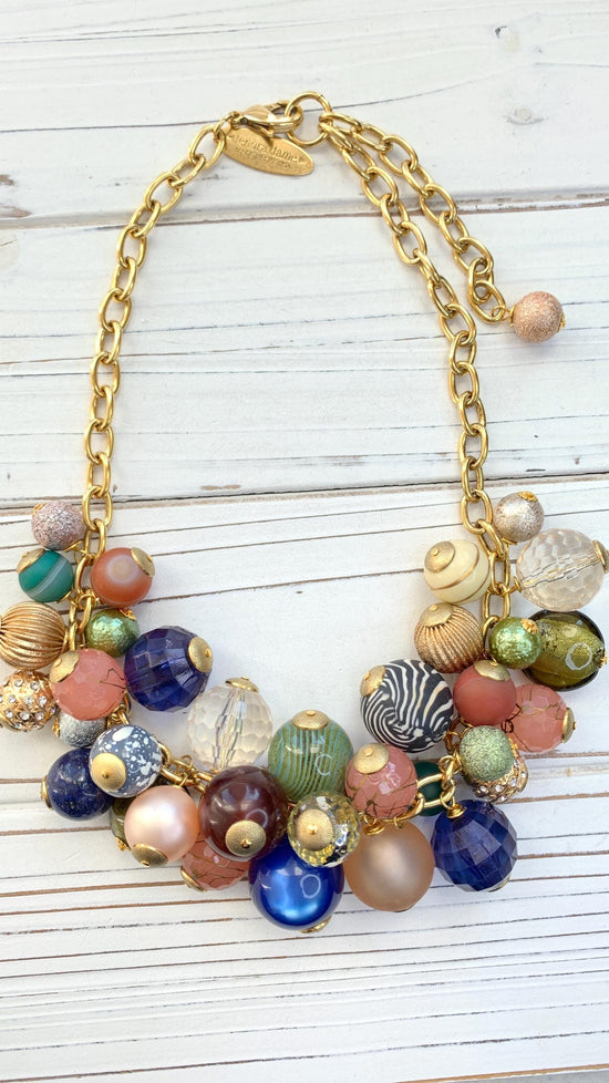 Gold chain necklace with multicolored beads.  The beads are various shapes and sizes, including round, faceted, and textured. Colors include pink, peach, green, blue, brown, and clear. Some beads have gold accents.  The necklace has a gold clasp with a small tag.  The beads appear to be glass or resin.