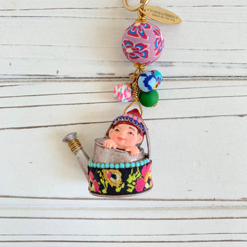 May include: A whimsical charm featuring a small figurine of a child sitting inside a silver watering can with a colorful floral design. The charm is attached to a gold chain with a pink, blue, and green patterned bead and a tag that reads &#39;Lenora Dame&#39; Made in the USA.