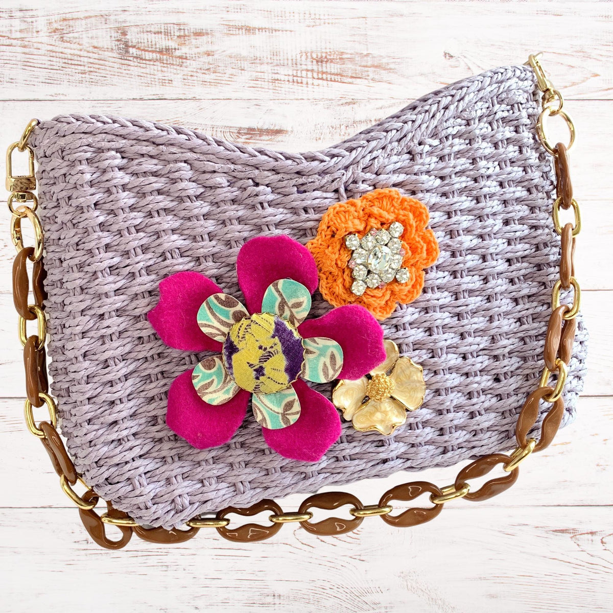 A lavender woven straw handbag with a brown chain strap and floral embellishments. The flowers are pink, orange, and yellow. The bag has a woven texture. Cute woven straw shoulder bag for summer.