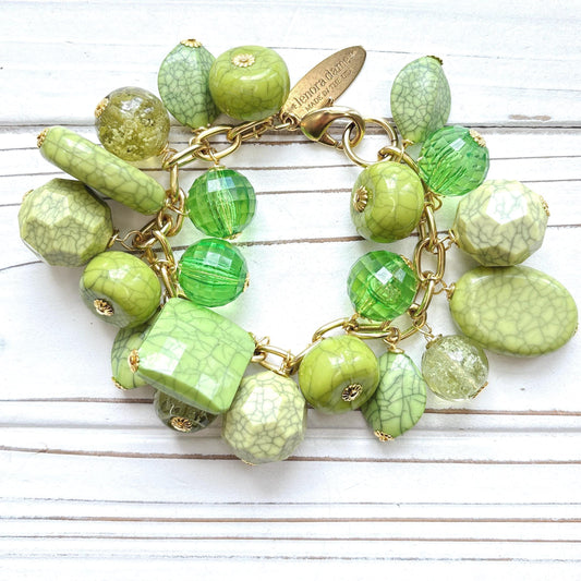 Gold chain charm bracelet filled with assorted green beads, some beads have a crackle look to them. Bracelet has a hangtag that says Lenora Dame, Handmade in the USA.