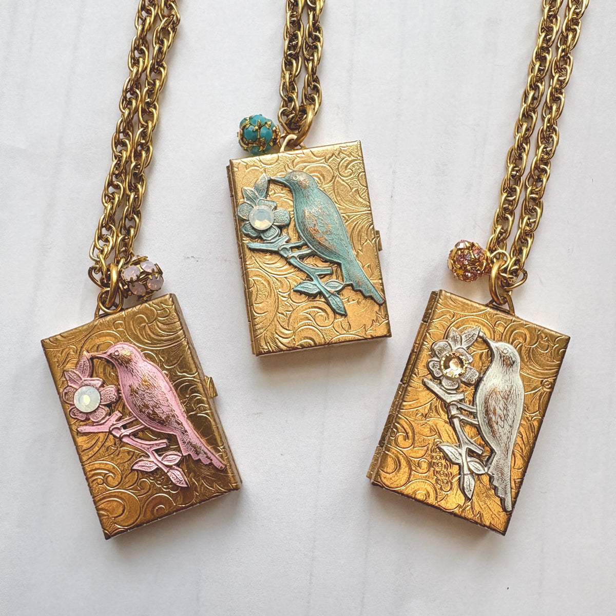 Three Brocade etched gold photo locket necklaces with gold rectangle lockets mounted with a metal bird on a flower stem design with rhinestone detail on the flower hung on a gold chain with a rhinestone bead ball. One locket has a pink bird design, the locket in the middle has blue bird design, and the locket on the right has a white bird design. Gold locket for women. Lenora Dame necklace made in the USA.