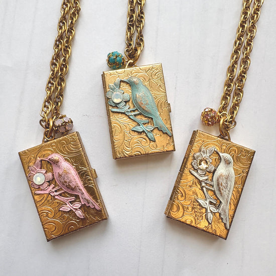 Three Brocade etched gold photo locket necklaces with gold rectangle lockets mounted with a metal bird on a flower stem design with rhinestone detail on the flower hung on a gold chain with a rhinestone bead ball. One locket has a pink bird design, the locket in the middle has blue bird design, and the locket on the right has a white bird design. Gold locket for women. Lenora Dame necklace made in the USA.