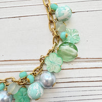 A mix of light green and light blue beads, pearls, and mother of pearl flowers hand from a gold chain necklace shown on a white washed wooden board. Green beaded necklace for women made in the USA. Lenora Dame necklace.