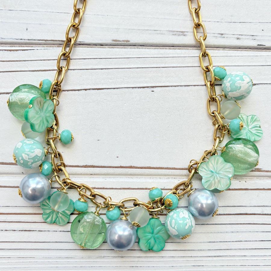 A mix of light green and light blue beads, pearls, and mother of pearl flowers hand from a gold chain necklace shown on a white washed wooden board. Green beaded necklace for women made in the USA. Lenora Dame necklace.