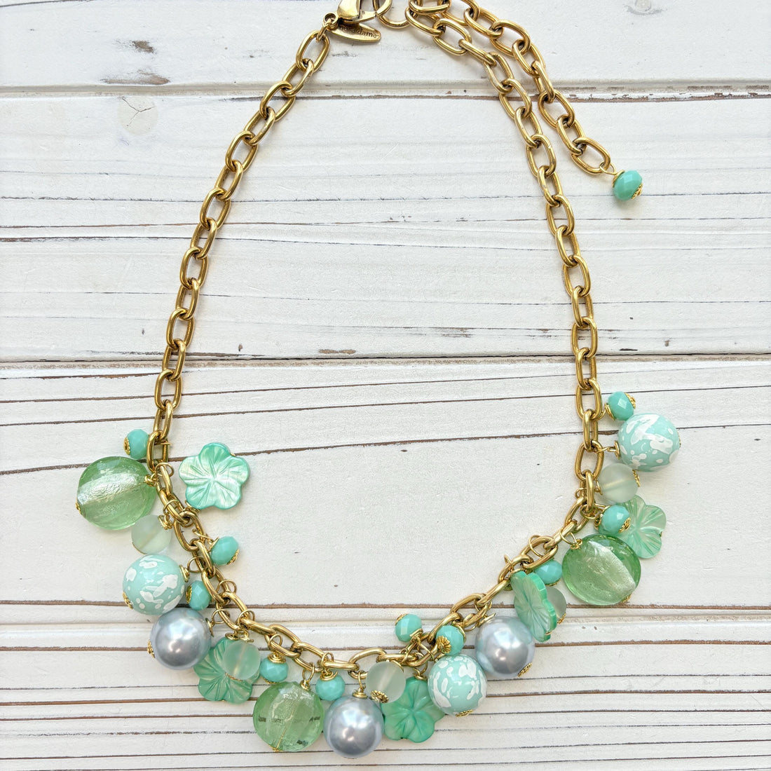 A mix of light green and light blue beads, pearls, and mother of pearl flowers hand from a gold chain necklace shown on a white washed wooden board. Green beaded necklace for women made in the USA. Lenora Dame necklace.