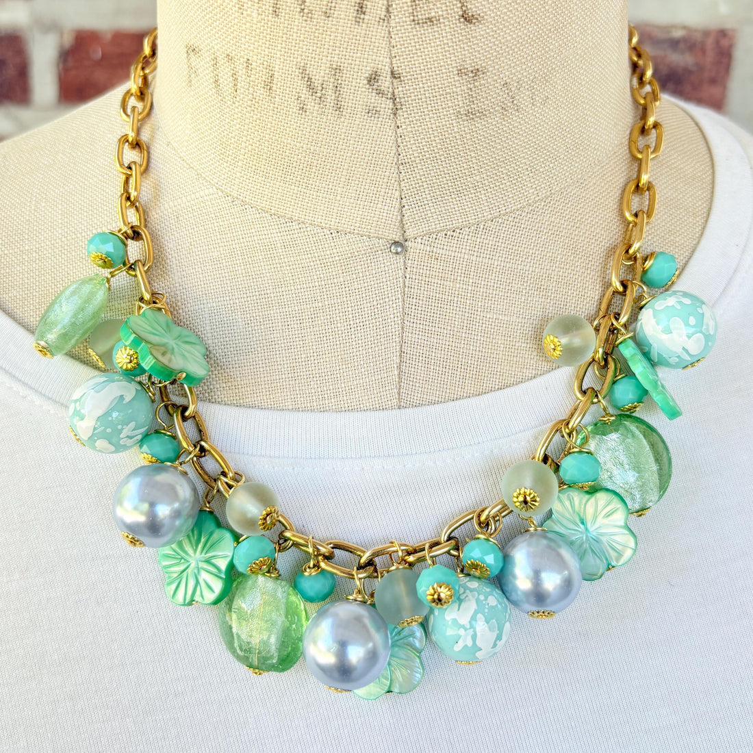A mix of light green and light blue beads, pearls, and mother of pearl flowers hand from a gold chain necklace shown on a dress form wearing a white shirt. Green beaded necklace for women made in the USA. Lenora Dame necklace.