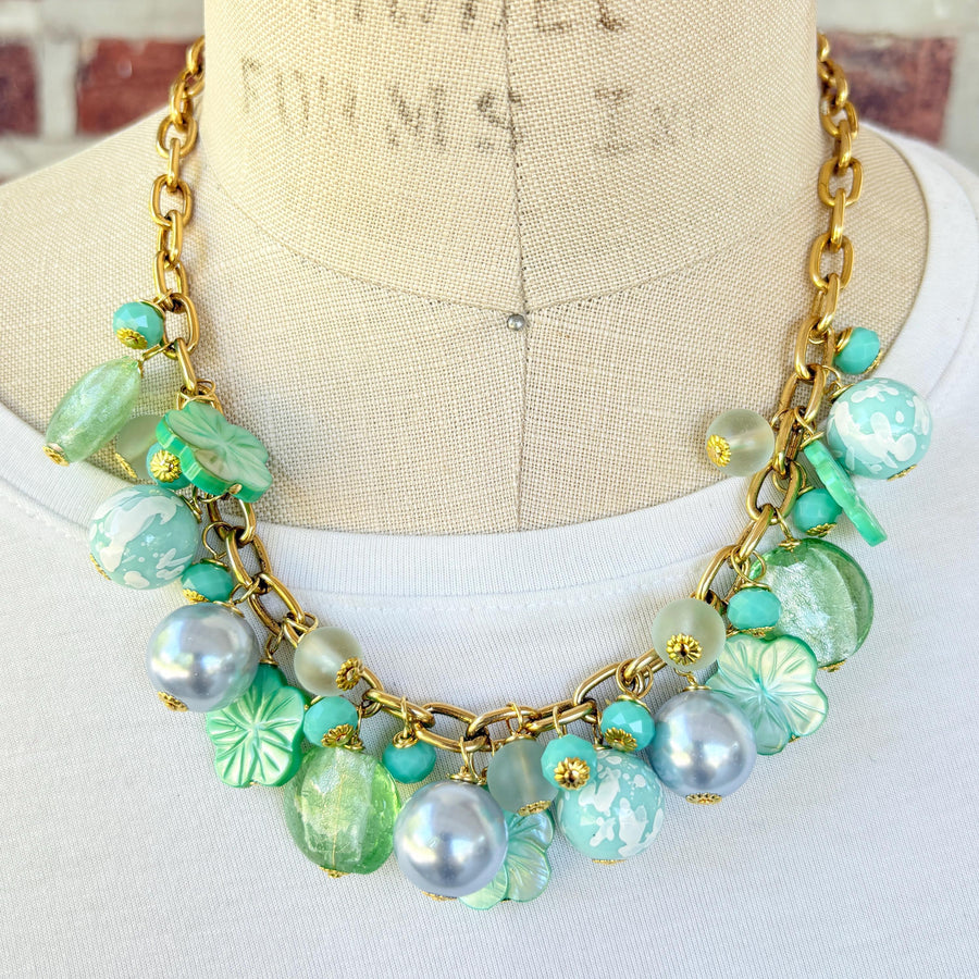 A mix of light green and light blue beads, pearls, and mother of pearl flowers hand from a gold chain necklace shown on a dress form wearing a white shirt. Green beaded necklace for women made in the USA. Lenora Dame necklace.