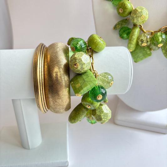 Gold chain charm bracelet filled with assorted green beads, some beads have a crackle look to them. Bracelet has a hangtag that says Lenora Dame, Handmade in the USA. Bracelet is shown on a white bracelet holder stacked with two gold bangle bracelets.
