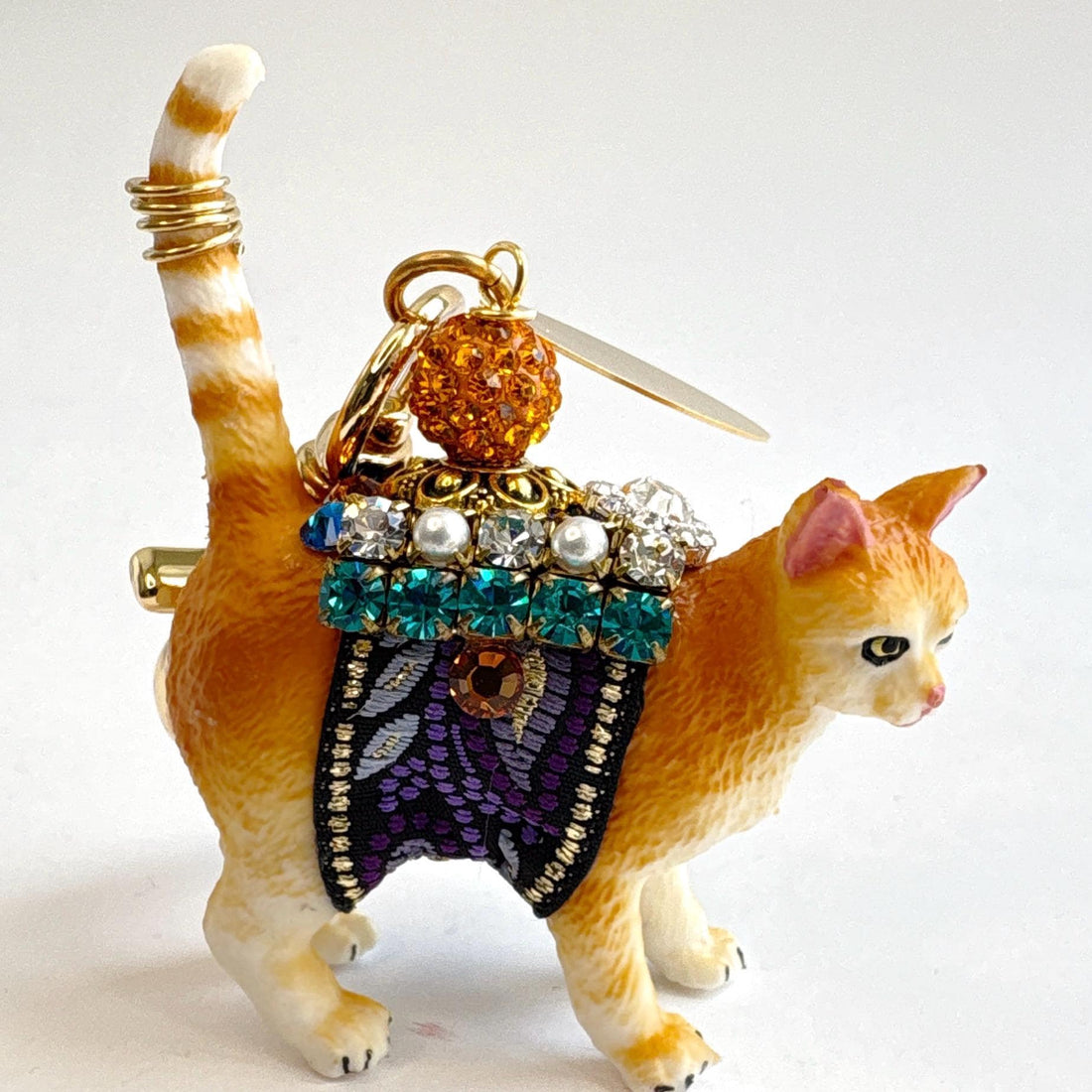 Cute cat bag charm with an orange tabby cat wearing purple and black ribbon trim embellished with green rhinestones, crystal rhinestones, and pearls. An orange rhinestone bead sits on the cat&