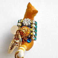 Cute cat bag charm with an orange tabby cat wearing purple and black ribbon trim embellished with green rhinestones, crystal rhinestones, and pearls. An orange rhinestone bead sits on the cat&#39;s back and is attached to the gold trigger clasp with Lenora Dame Made in USA hangtag. Lenora Dame bag charm.