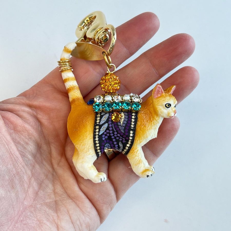 Cute cat bag charm with an orange tabby cat wearing purple and black ribbon trim embellished with green rhinestones, crystal rhinestones, and pearls shown being held in a human hand for size scale. An orange rhinestone bead sits on the cat&#39;s back and is attached to the gold trigger clasp with Lenora Dame Made in USA hangtag. Lenora Dame bag charm.