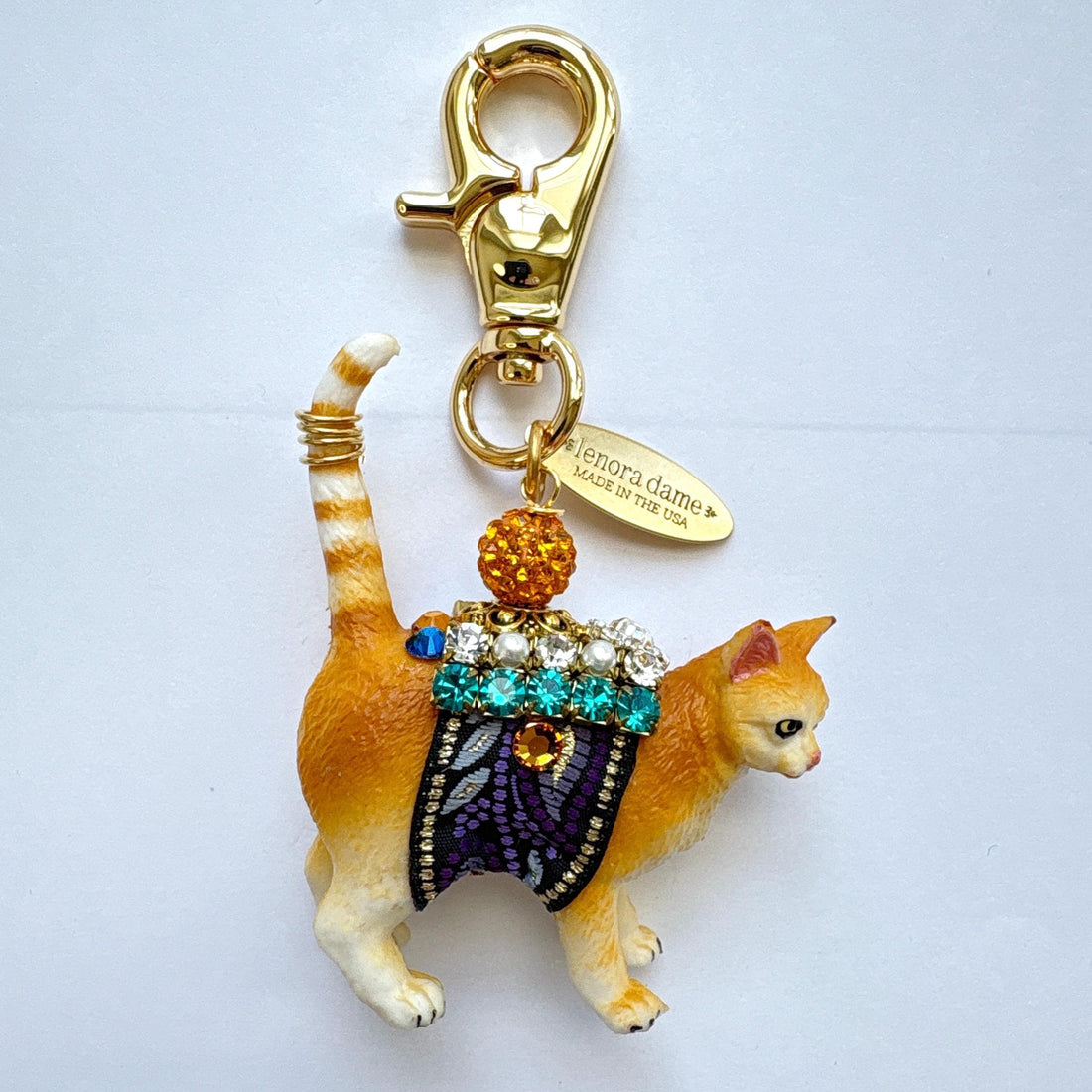 Cute cat bag charm with an orange tabby cat wearing purple and black ribbon trim embellished with green rhinestones, crystal rhinestones, and pearls. An orange rhinestone bead sits on the cat&