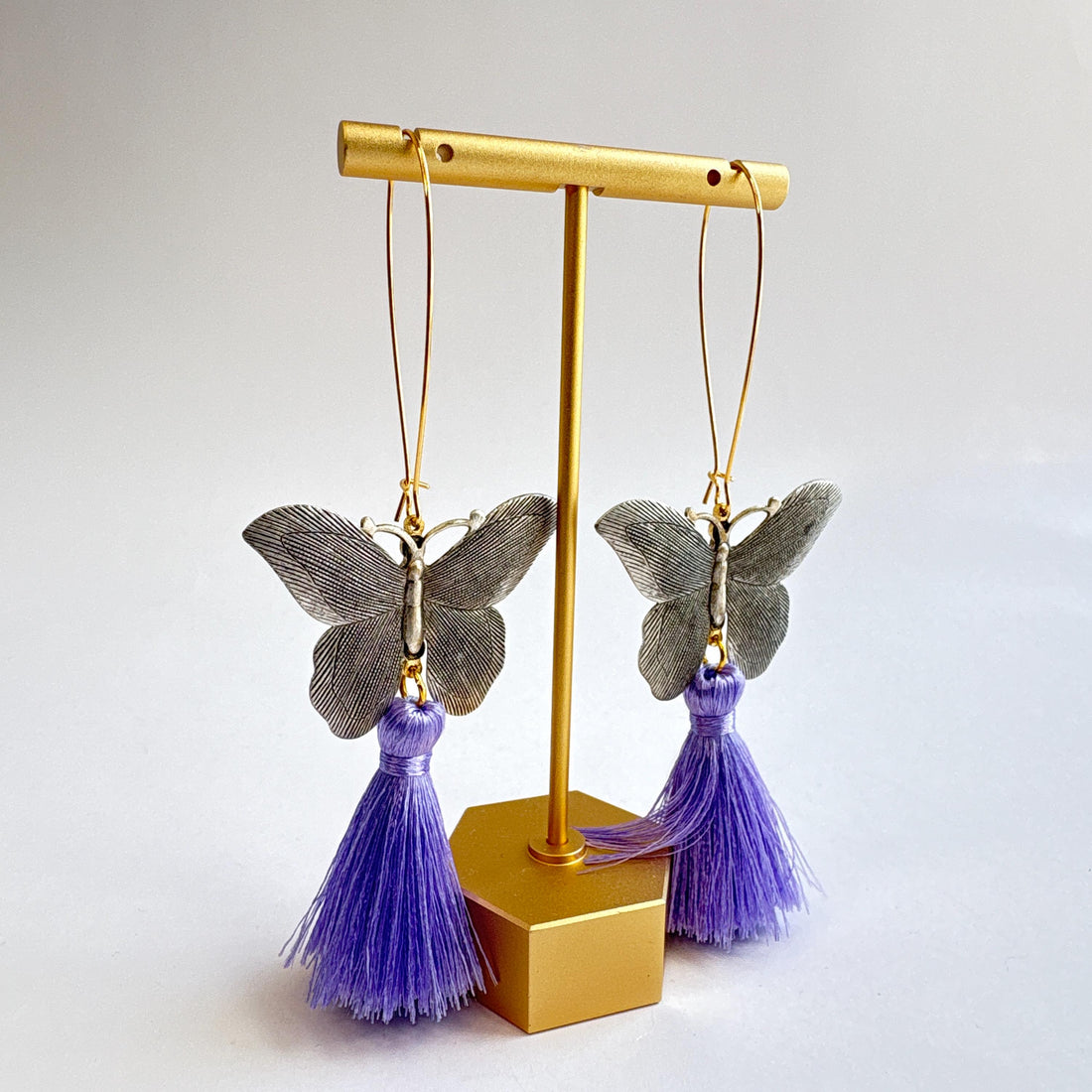 Butterfly dangle earrings with silver metal butterflies, hanging from long oval gold hoop ear wires. Light purple silky tassels hang from the bottom of the butterfly earrings. Lenora Dame Jewelry made in the USA.