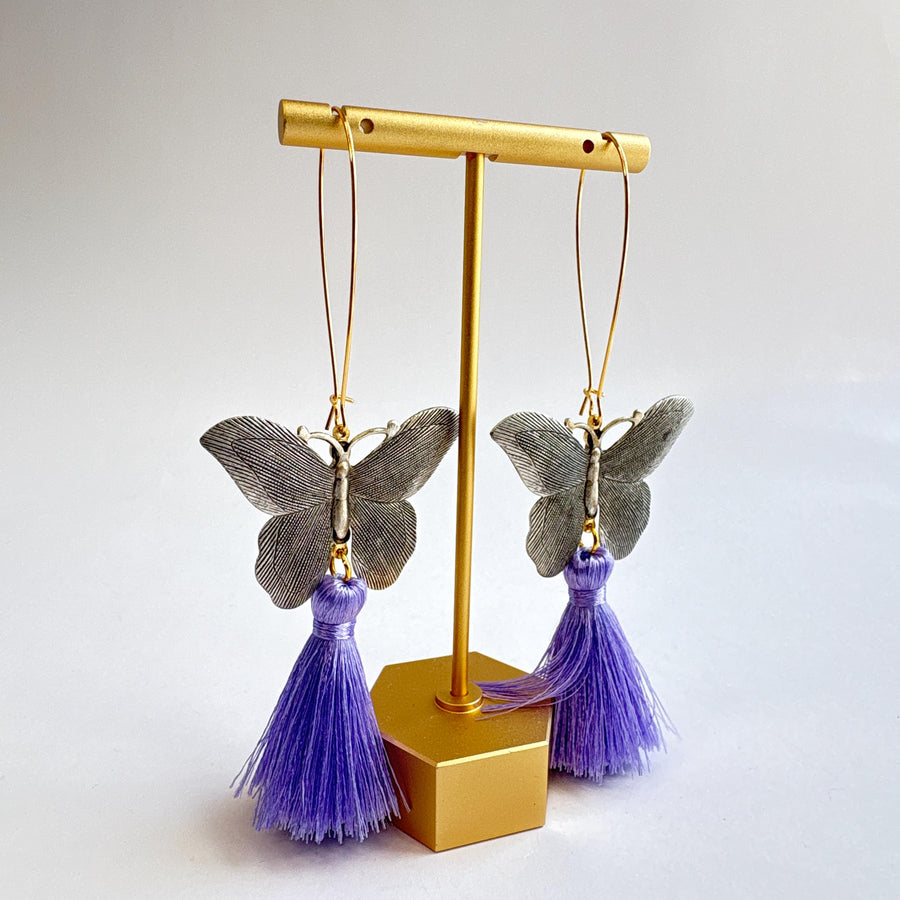 Butterfly dangle earrings with silver metal butterflies, hanging from long oval gold hoop ear wires. Light purple silky tassels hang from the bottom of the butterfly earrings. Lenora Dame Jewelry made in the USA.