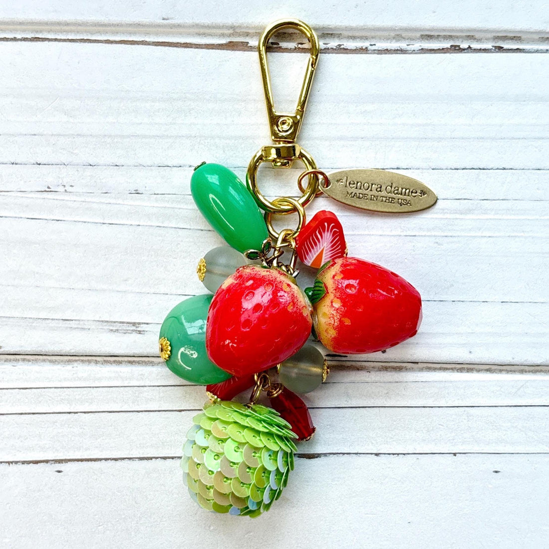 Cute strawberry bag charm with a mix of red and green beads, two red strawberry charms on a gold chain with gold trigger clasp and brass hang tag that says Lenora Dame Made in USA. Lenora Dame bag charm.