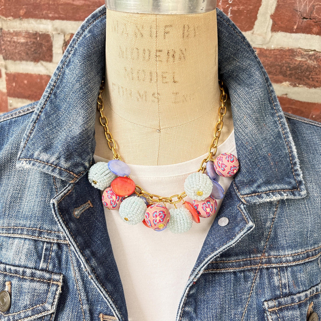 Chunky charm necklace with assorted mix of light blue seed bead balls, floral fimo beads, blue and orange mother of pearl disc bead hung on a gold necklace chain with lobster clasp and hangtag that says Lenora Dame Made in the USA.  Necklace is shown on a dress form wearing a white shirt and blue denim jacket with a red brick wall background. Lenora Dame necklace.