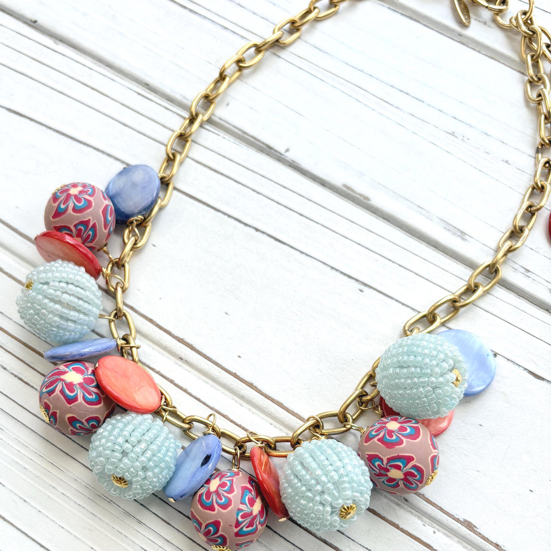 Chunky charm necklace with assorted mix of light blue seed bead balls, floral fimo beads, blue and orange mother of pearl disc bead hung on a gold necklace chain with lobster clasp and hangtag that says Lenora Dame Made in the USA. Lenora Dame necklace.