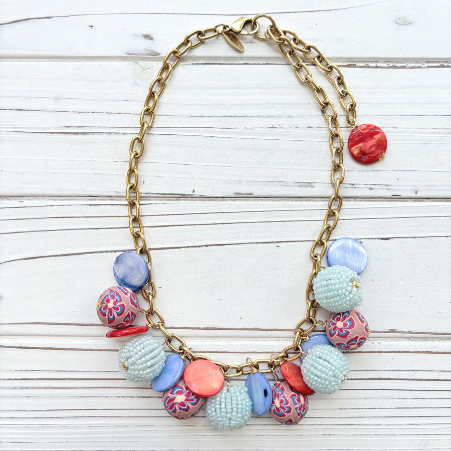 Chunky charm necklace with assorted mix of light blue seed bead balls, floral fimo beads, blue and orange mother of pearl disc bead hung on a gold necklace chain with lobster clasp and hangtag that says Lenora Dame Made in the USA. Lenora Dame necklace.