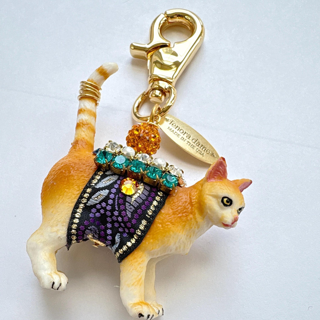 Cute cat bag charm with an orange tabby cat wearing purple and black ribbon trim embellished with green rhinestones, crystal rhinestones, and pearls. An orange rhinestone bead sits on the cat&
