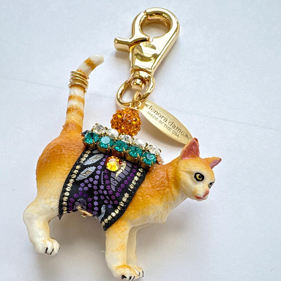Cute cat bag charm with an orange tabby cat wearing purple and black ribbon trim embellished with green rhinestones, crystal rhinestones, and pearls. An orange rhinestone bead sits on the cat&#39;s back and is attached to the gold trigger clasp with Lenora Dame Made in USA hangtag. Lenora Dame bag charm.