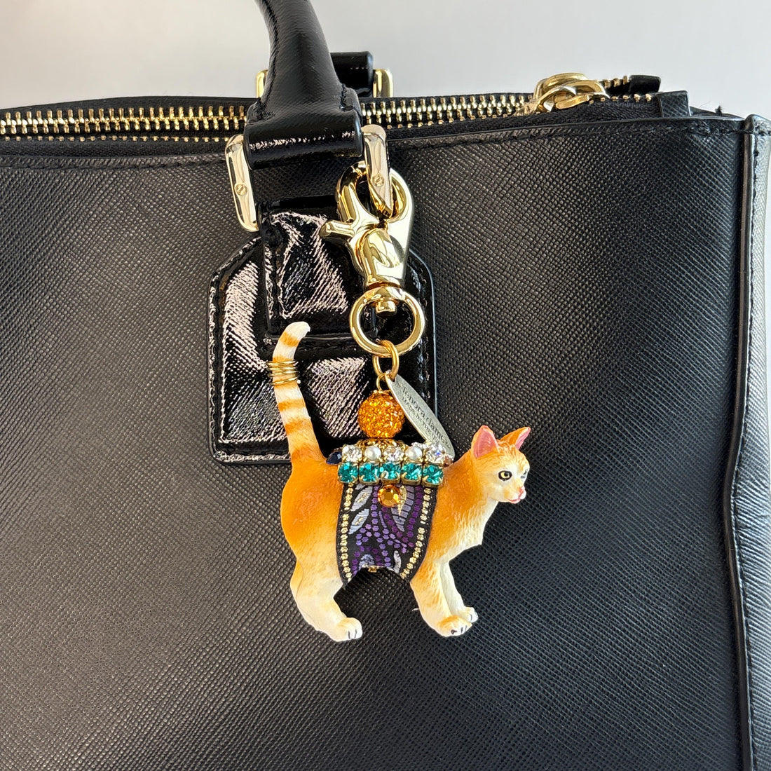 Cute cat bag charm with an orange tabby cat wearing purple and black ribbon trim embellished with green rhinestones, crystal rhinestones, and pearls shown attached to the strap of a black handbag. An orange rhinestone bead sits on the cat&