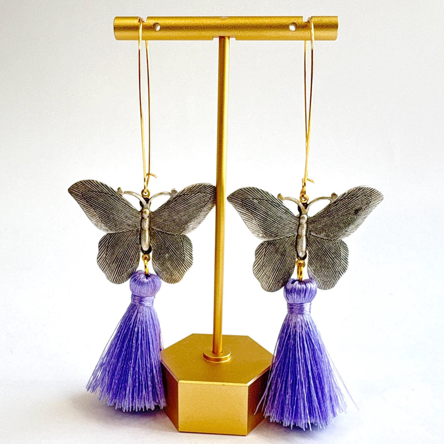 Butterfly dangle earrings with silver metal butterflies, hanging from long oval gold hoop ear wires. Light purple silky tassels hang from the bottom of the butterfly earrings. Lenora Dame Jewelry made in the USA.