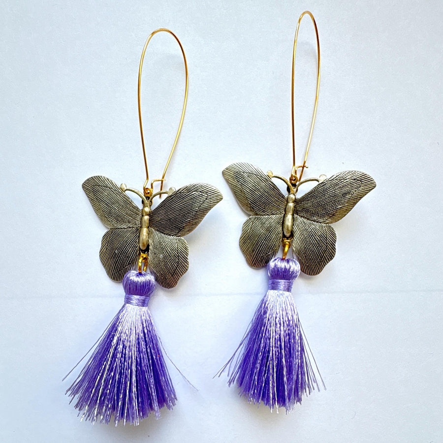 Butterfly dangle earrings with silver metal butterflies, hanging from long oval gold hoop ear wires. Light purple silky tassels hang from the bottom of the butterfly earrings. Lenora Dame Jewelry made in the USA.