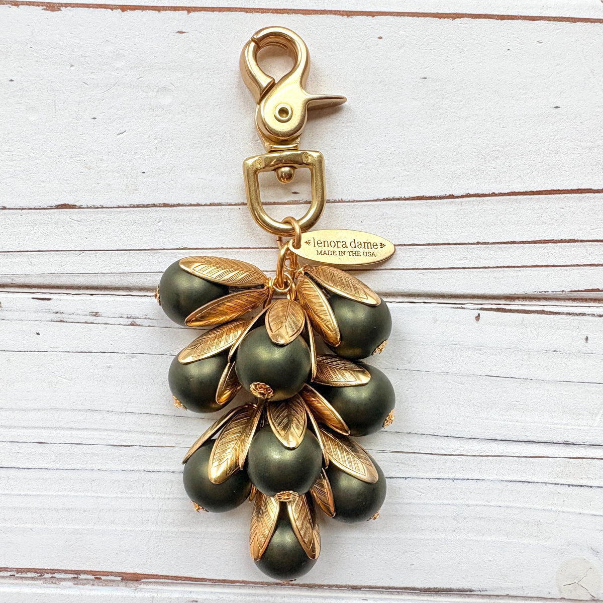 Gold bag charm with a trigger clasp at the top holding a cluster of matte hunter green pearls encased in gold leaf bead caps. A gold oval tag imprinted with Lenora Dame Handmade in USA hangs at the top by the trigger clasp. Lenora Dame bag charms. Green beaded purse charm.