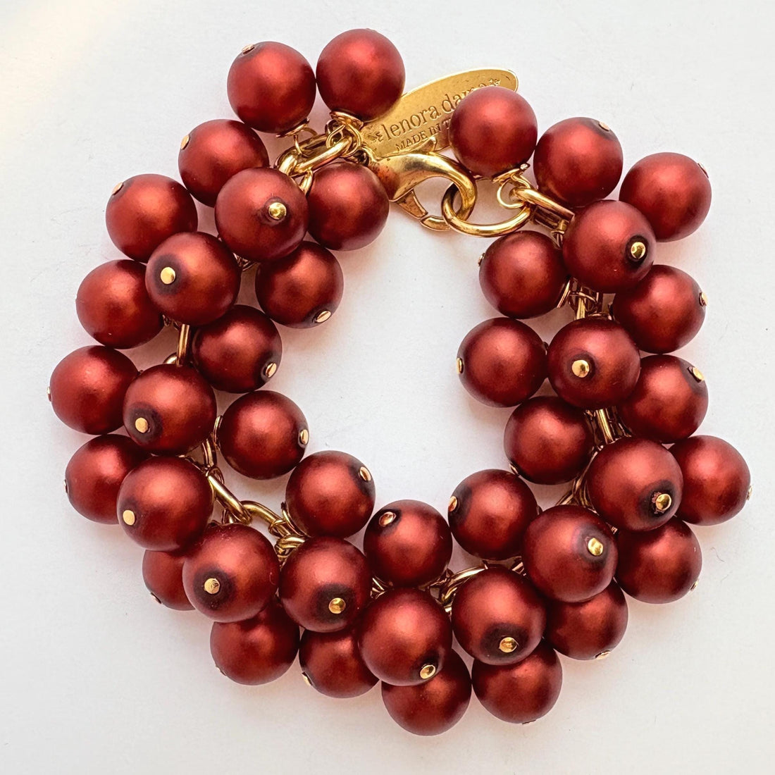 A gold chain bracelet with multiple strands of matte dark red faux pearls. The bracelet has a small tag that reads &