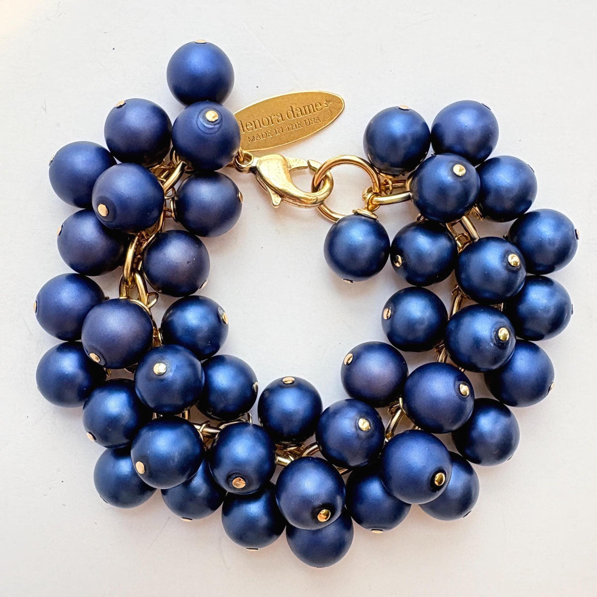 A gold chain bracelet with multiple strands of matte navy blue faux pearls. The bracelet has a small tag that reads 'Lenora Dame' and 'Made in the USA.'
