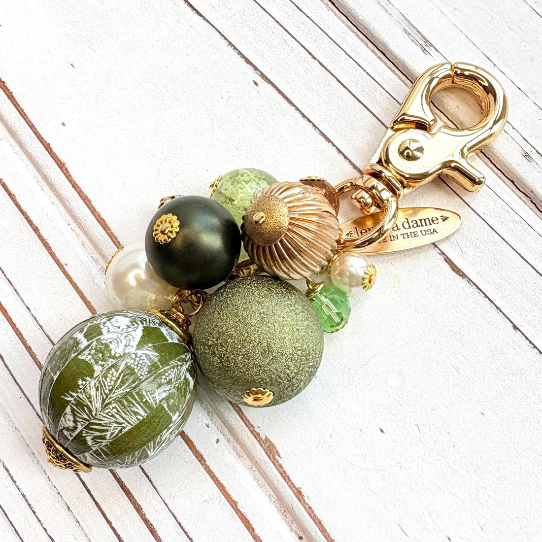 Beaded bag charm with a mix of bead styles in shades of green, gold, and pearl. One larger round bead in a army green and white toile print hangs at the bottom and a gold trigger clasp on top of all of the beads. Brass oval hang tag near the trigger clasp that says &quot;Lenora Dame Handmade in USA&quot;. Lenora Dame bag charm.