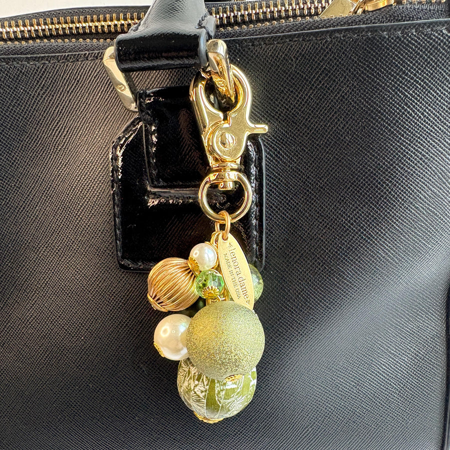 Beaded bag charm with a mix of bead styles in shades of green, gold, and pearl. One larger round bead in a army green and white toile print hangs at the bottom and a gold trigger clasp on top of all of the beads. Brass oval hang tag near the trigger clasp that says &quot;Lenora Dame Handmade in USA&quot;. Lenora Dame bag charm.