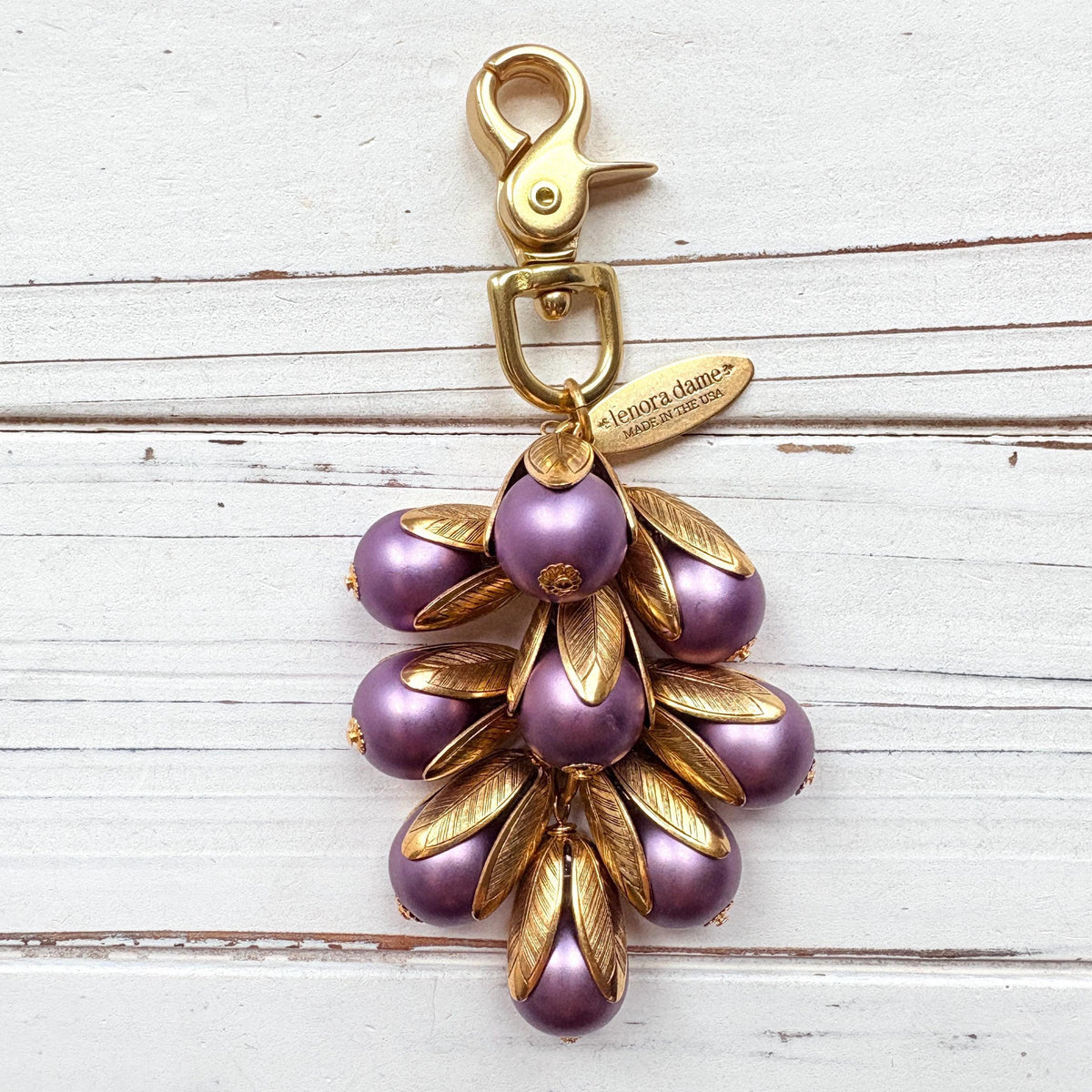 Gold bag charm with a trigger clasp at the top holding a cluster of lavender matte purple pearls encased in gold leaf bead caps. A gold oval tag imprinted with Lenora Dame Handmade in USA hangs at the top by the trigger clasp. Lenora Dame bag charms. Purple purse charm.