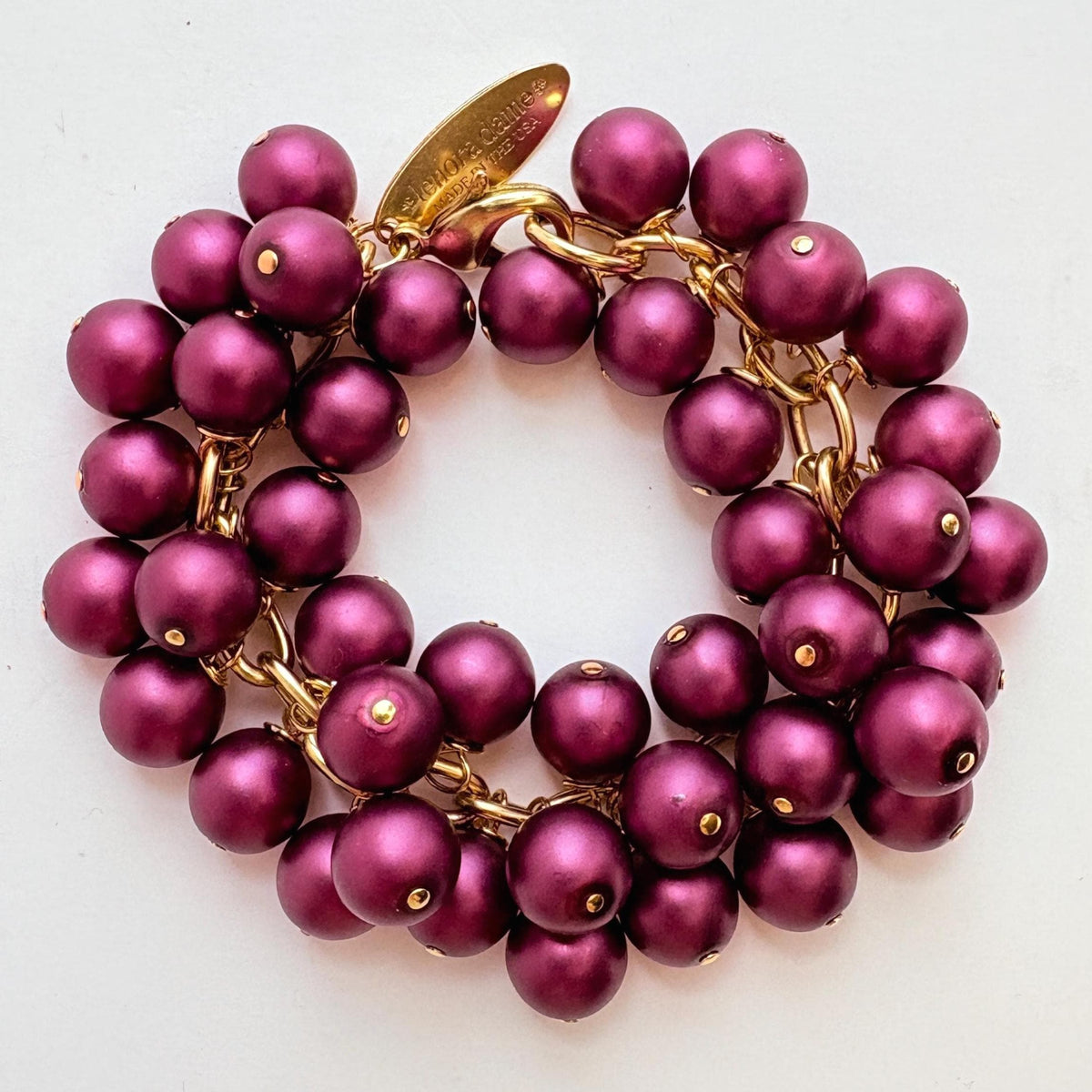 A gold chain bracelet with multiple strands of dark purple faux pearls. The bracelet has a small tag that reads 'Lenora Dame' and 'Made in the USA.'