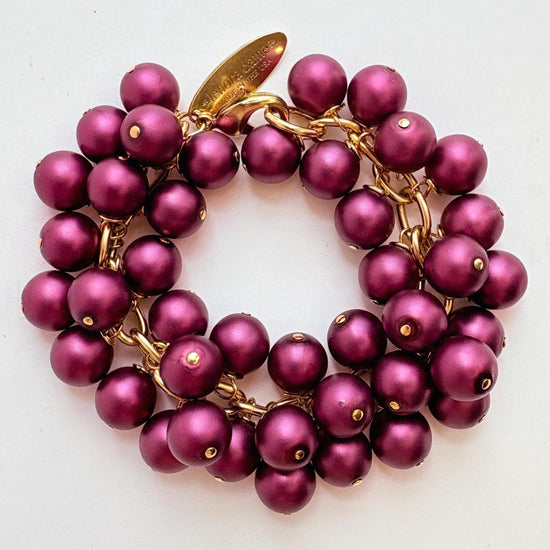 A gold chain bracelet with multiple strands of dark purple faux pearls. The bracelet has a small tag that reads 'Lenora Dame' and 'Made in the USA.'