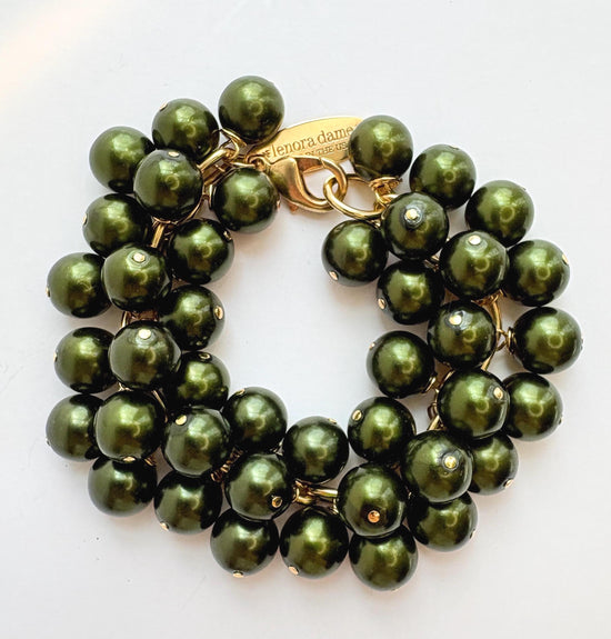 A gold chain bracelet with multiple strands of shiny hunter green faux pearls. The bracelet has a small tag that reads 'Lenora Dame' and 'Made in the USA.'