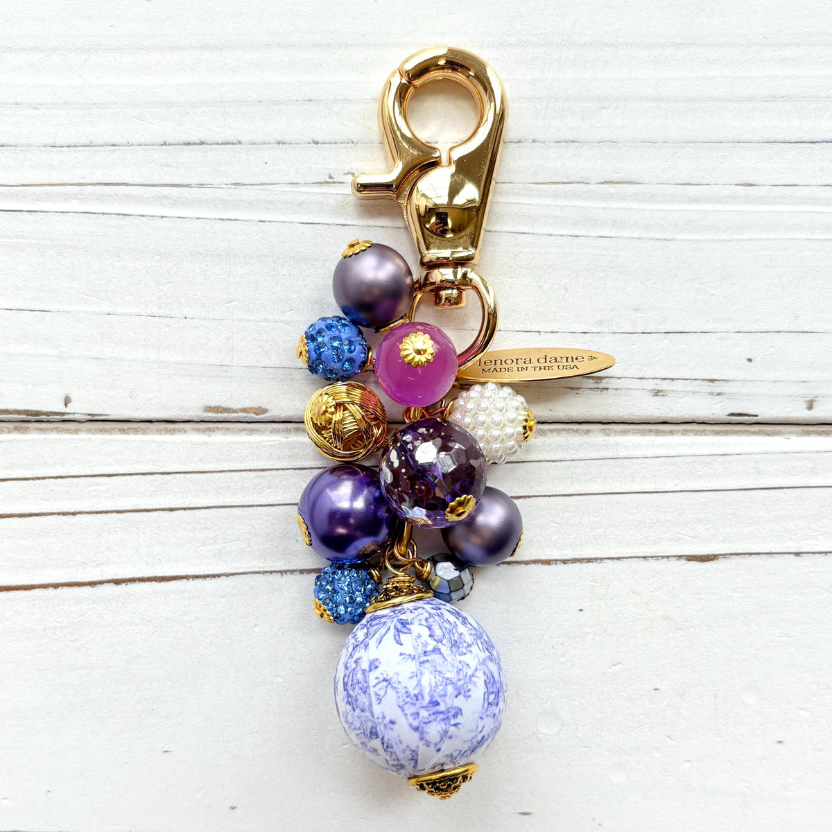 Beaded bag charm with a mix of bead styles in shades of purple, blue, gold and pearl. One larger round bead in a purple and white toile print hangs at the bottom and a gold trigger clasp on top of all of the beads. Brass oval hang tag near the trigger clasp that says "Lenora Dame Handmade in USA". Lenora Dame bag charm.