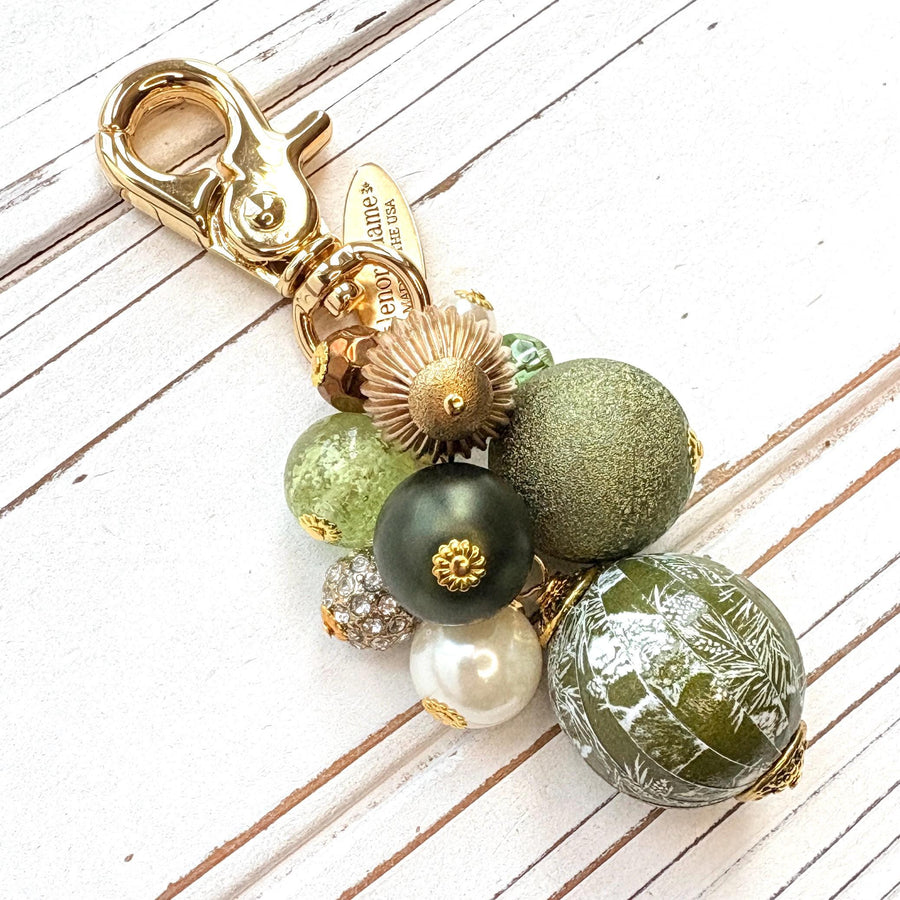 Beaded bag charm with a mix of bead styles in shades of green, gold, and pearl. One larger round bead in a army green and white toile print hangs at the bottom and a gold trigger clasp on top of all of the beads. Brass oval hang tag near the trigger clasp that says &quot;Lenora Dame Handmade in USA&quot;. Lenora Dame bag charm.
