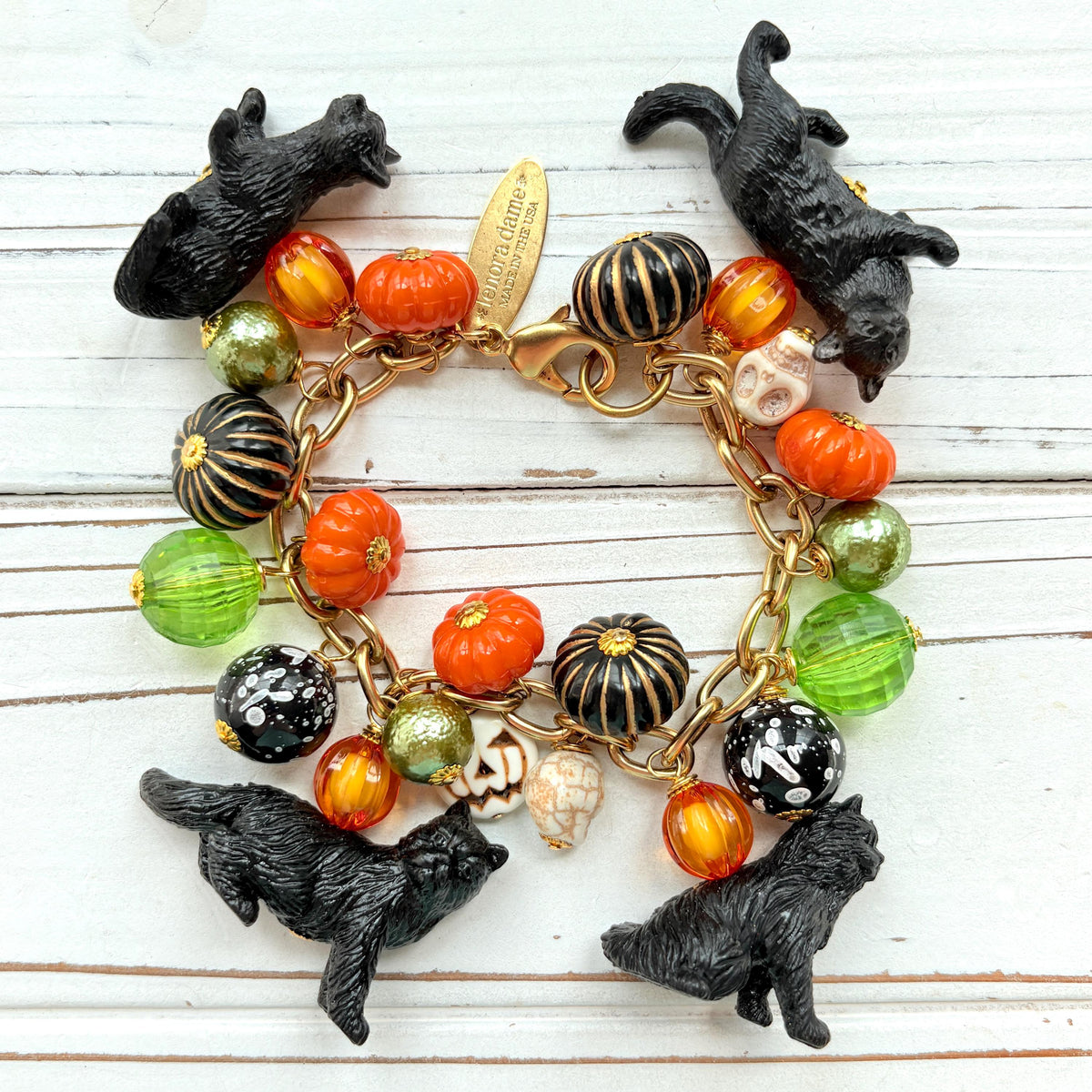 A gold chain bracelet with black cat charms, green and orange beads, and a white pumpkin charm. The bracelet has a tag that reads 'Lenora Dame' and 'Made in USA'.