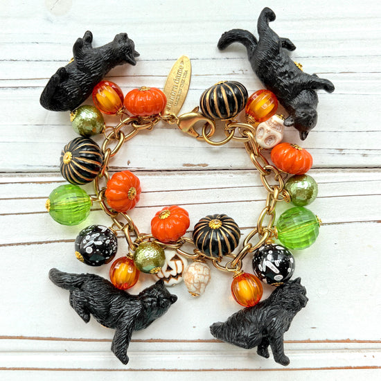 A gold chain bracelet with black cat charms, green and orange beads, and a white pumpkin charm. The bracelet has a tag that reads 'Lenora Dame' and 'Made in USA'.