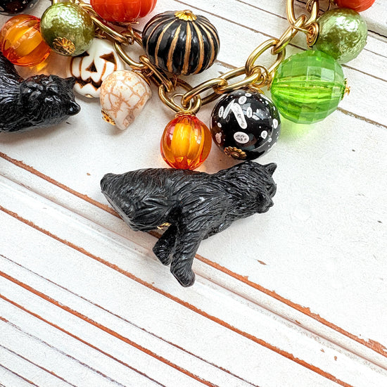A gold chain bracelet with black cat charms, green and orange beads, and a white pumpkin charm. The bracelet has a tag that reads 'Lenora Dame' and 'Made in USA'.