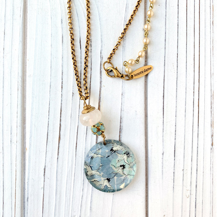 May include: A gold chain necklace with a round glass pendant featuring a blue and white pattern of flying birds. The pendant is attached to a white bead and a cluster of smaller blue beads. The necklace has a gold clasp and a small gold tag that reads &#39;lenora dame&#39;.