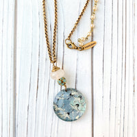 May include: A gold chain necklace with a round glass pendant featuring a blue and white pattern of flying birds. The pendant is attached to a white bead and a cluster of smaller blue beads. The necklace has a gold clasp and a small gold tag that reads &#39;lenora dame&#39;.