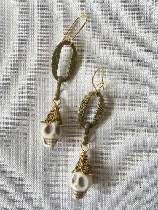 Lenora Dame Shackled Skull Earrings with white carved sugar skulls topped with brass filigree cone bead dangling from oval etched metal pieces in an antique bronze finish. Earrings hang from gold French hook ear wires.
