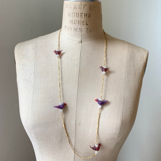 Little purple glass lampworked birds spaced out between bits of gold plated bar chain to create a long necklace. Shown on a vintage dress making form.
