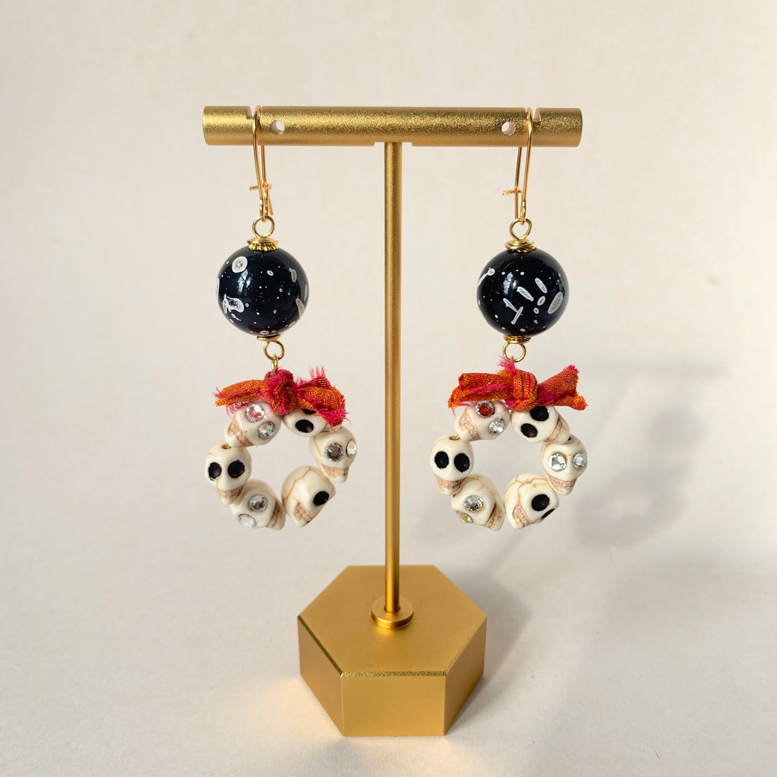 Lenora Dame Halloween earrings with white miniature carved sugar skulls, some with rhinestone embellished eyes, shaped into hoops and adorned with a bow made from orange and red sari silk. The skull hoops dangle from black with white splatter acrylic beads attached to gold-plated French hook ear wires.